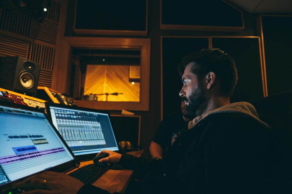 Side view of concentrated ethnic bearded soundman working on computers with musical program on screens while creating arrangement sitting at table near colleague in music studio