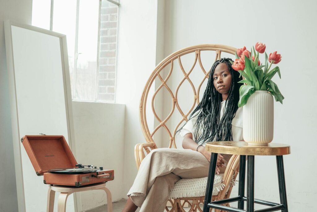 Ethnic woman sitting in armchair near vinyl record player and vase of fresh flowers